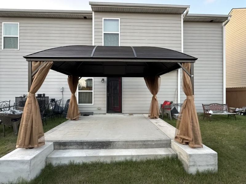 Front view of the same hardtop gazebo, with tan curtains tied back at each post on the raised concrete patio