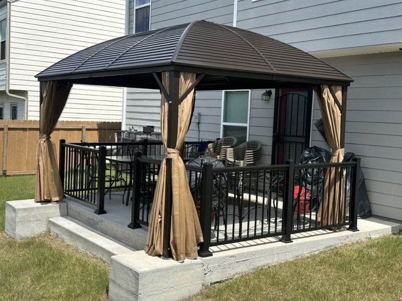 Hardtop gazebo with a dark curved metal roof and tan curtains on a raised concrete patio with black railings, beside a two-story house