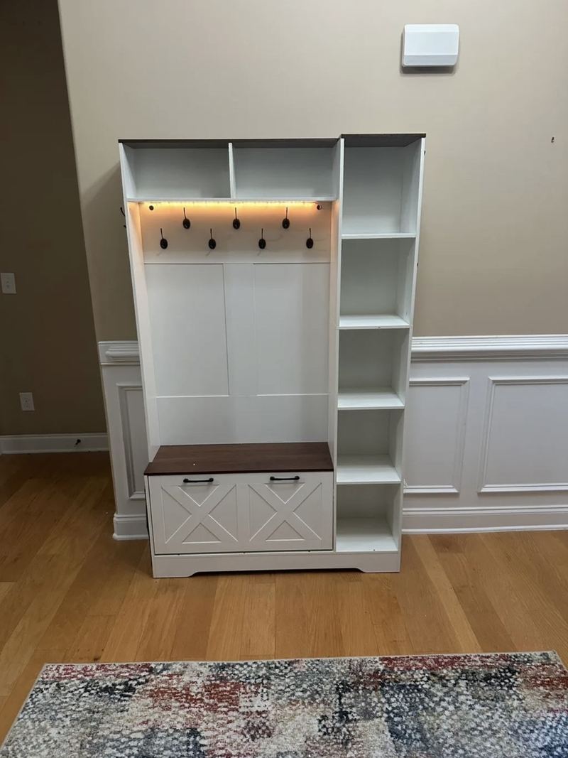 White hall tree with open cubbies, black coat hooks under an LED light strip, a wood-top storage bench and a side shelving tower