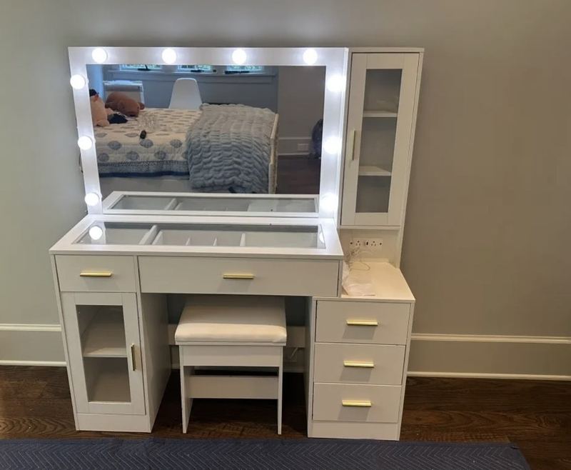 White vanity with a lighted mirror, glass-front cabinets, drawers with gold pulls and a matching stool